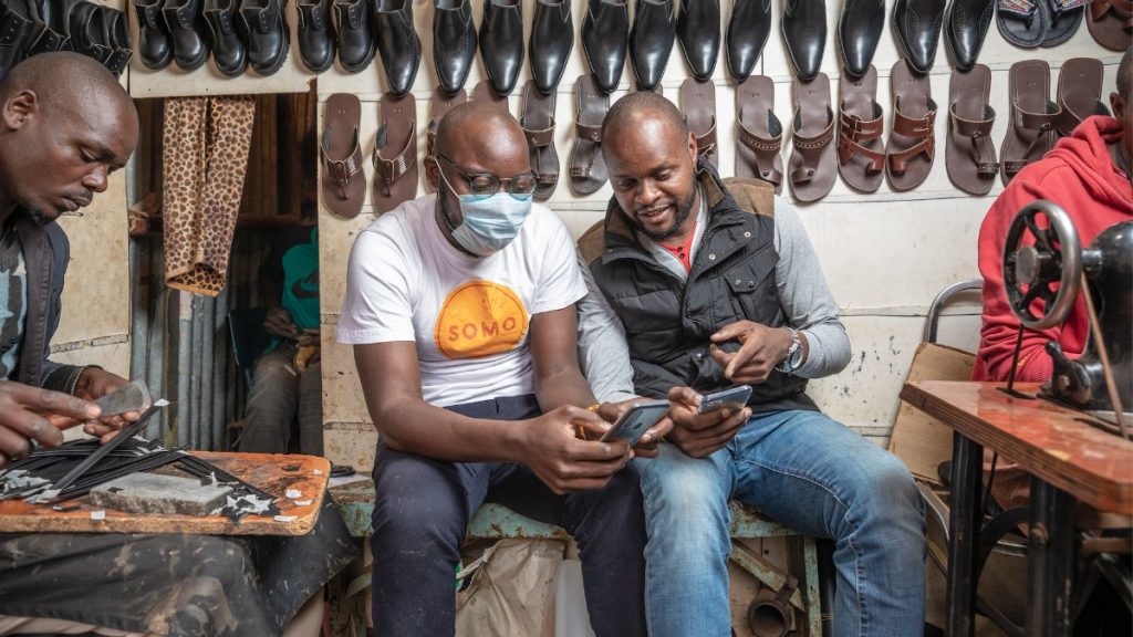 Four men in a shoe making shop; two working on shoes and two others looking at something on a phone